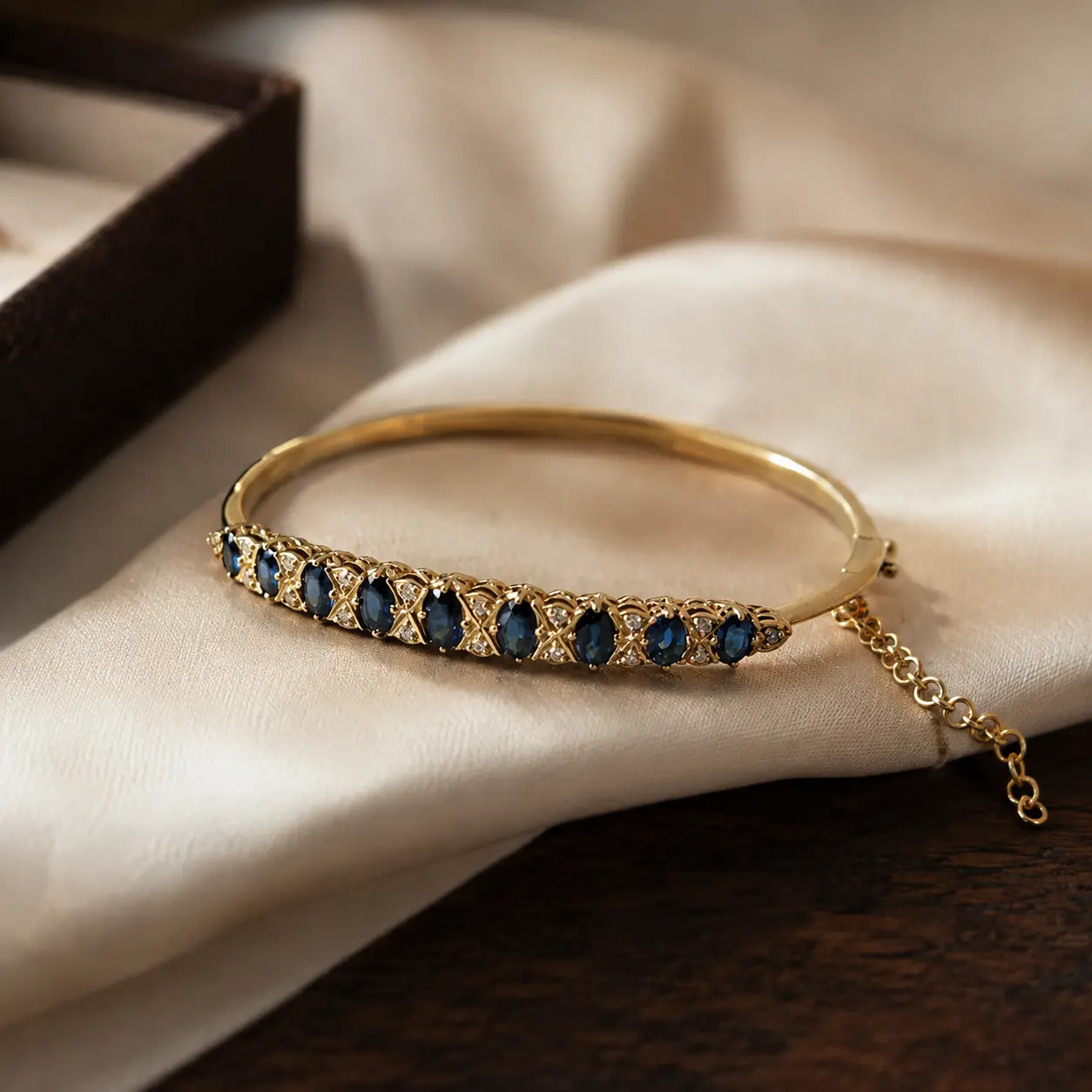 A yellow gold hinged bangle set with a row of oval blue sapphires and small diamonds on cream silk